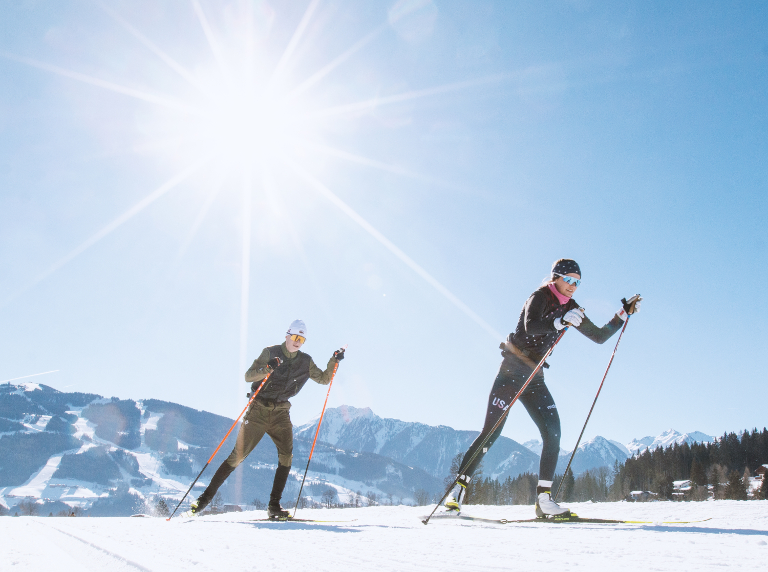 Das Bild zeigt zwei Personen beim Langlaufen auf einer verschneiten Fläche an einem sonnigen Tag.Die beiden Langläufer tragen sportliche Kleidung und Skistöcke. Im Hintergrund ist eine bergige Winterlandschaft mit Pisten und Wäldern zu sehen.