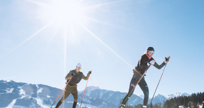 Langlauf Hotel Ramsau Das Bild zeigt zwei Personen beim Langlaufen auf einer verschneiten Fläche an einem sonnigen Tag.Die beiden Langläufer tragen sportliche Kleidung und Skistöcke. Im Hintergrund ist eine bergige Winterlandschaft mit Pisten und Wäldern zu sehen.