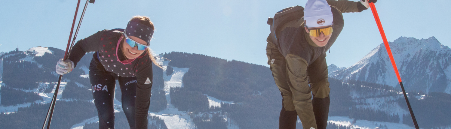 Das Bild zeigt zwei lachende Langläuferinen. Sie haben beide in einer Hand die Langlaufstöcke, mit der anderen berühren sie die Bindung des Skis. Sie sind in einer gebückten Haltung. Im Hintergrund erstreckt sich eine atemberaubende Alpinlandschaft.