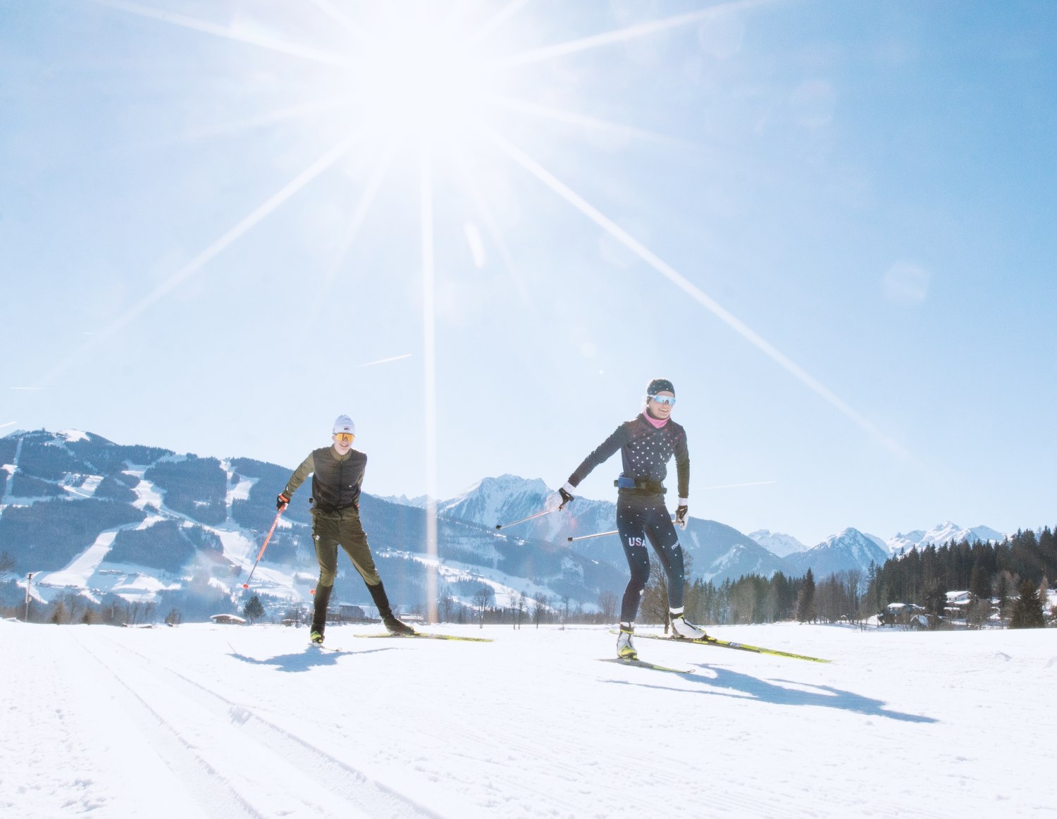 Das Bild zeigt zwei Personen beim Langlaufen auf einer verschneiten Fläche an einem sonnigen Tag.Die beiden Langläufer tragen sportliche Kleidung und Skistöcke. Im Hintergrund ist eine bergige Winterlandschaft mit Pisten und Wäldern zu sehen.