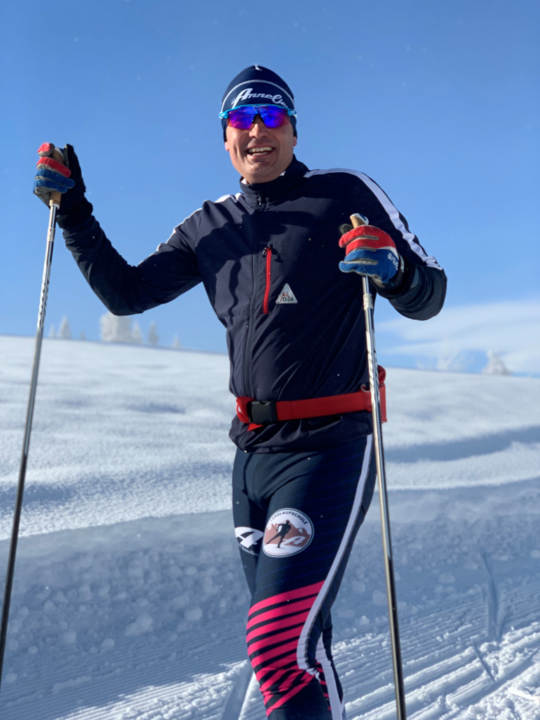 Roland - Langlaufen Das Bild zeigt einen lächelnden Mann in Langlaufausrüstung, der mit Langlaufstöcken im Schnee steht. Er trägt eine blaue Mütze, eine dunkle Jacke sowie eine dunkle Hose mit pink-weißen Streifen. Eine rote Bauchtasche ist um seine Hüfte geschnallt. Er trägt eine Sonnenbrille. Im Hintergrund ist eine verschneite Landschaft unter blauem Himmel zu sehen.