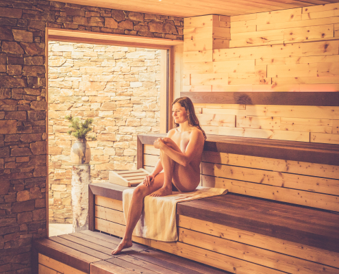 Das Bild zeigt eine Frau, die in einer Sauna aus hellem Holz auf einer Bank sitzt. Sie hat ein Handtuch unter sich gelegt und wirkt entspannt. Durch eine Öffnung ist eine Wand aus Naturstein sichtbar.