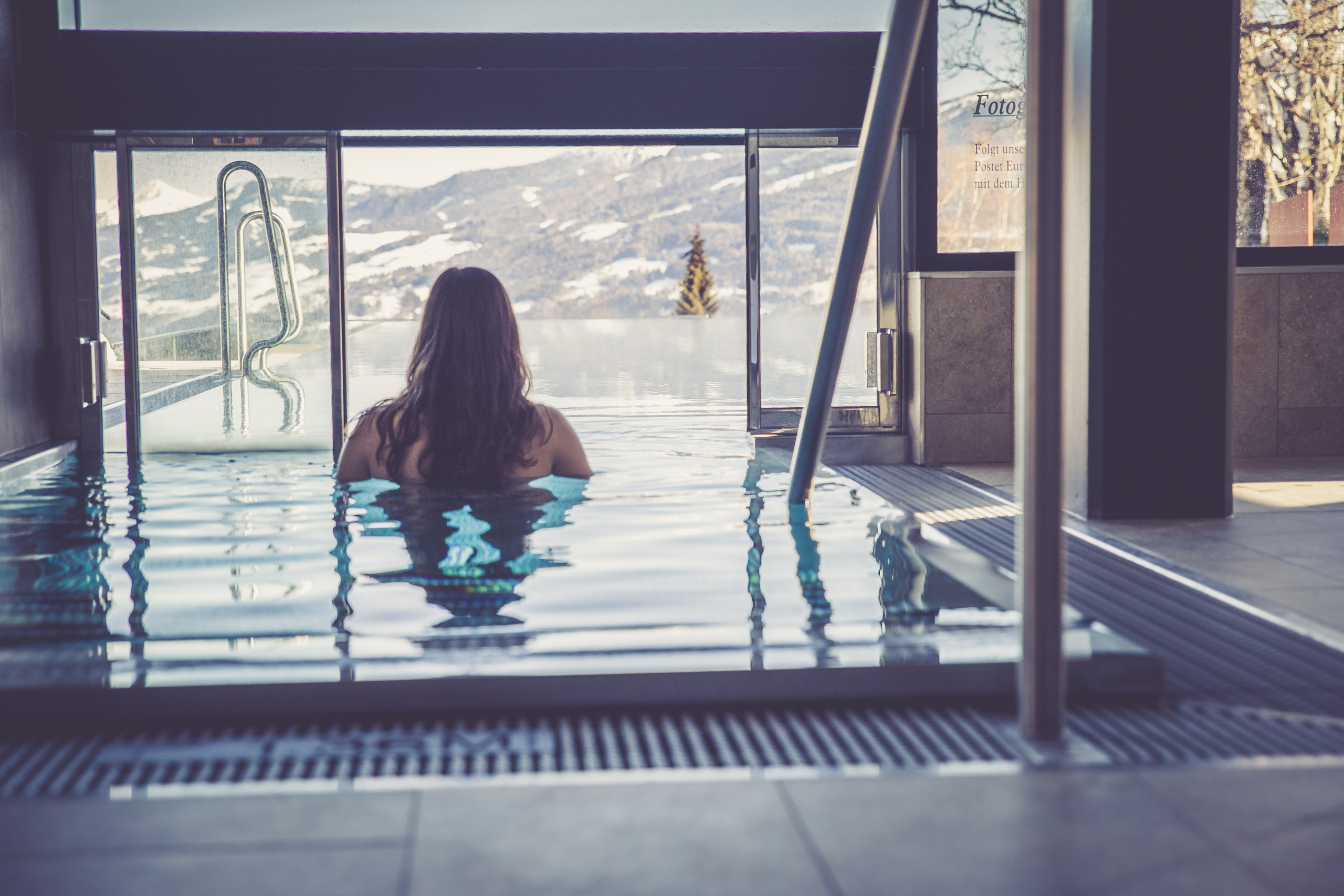 Wellness und Sauna Hotel Annelies inkl. Pool Im Fokus steht eine Frau, die sich im Innenbreich des Infinity-Pools befindet. Vor ihr eine geöffnete Fensterfront, die als Übergang vom Innen zum Außenbereich dient. Draußen ist ein Infinity-Pool zu sehen, der scheinbar in eine verschneite Berglandschaft übergeht.