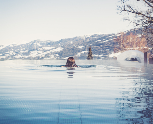 Das Bild zeigt eine Frau, die in einem Infinity-Pool schwimmt, mit Blick auf eine schneebedeckte Berglandschaft.