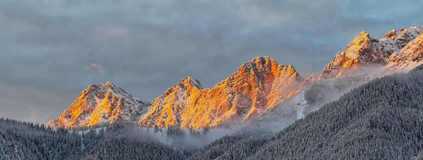 Schneebedeckter Dachstein bei Sonnenuntergang Berglandschaft im Winter bei Sonnenuntergang. Die Berggipfel sind teilweise schneebedeckt, wobei die Gipfel von der untergehenden Sonne golden beleuchtet werden. Im Vordergrund erstreckt sich ein verschneiter Wald.