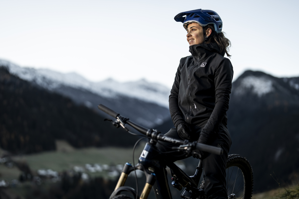 Eine Bikerin, mit blauem Helm, sitzt auf ihrem Bike und genießt die Aussicht. Eine beeindruckende Bergfront sowie ein kleines Tal bilden den Hintergund.