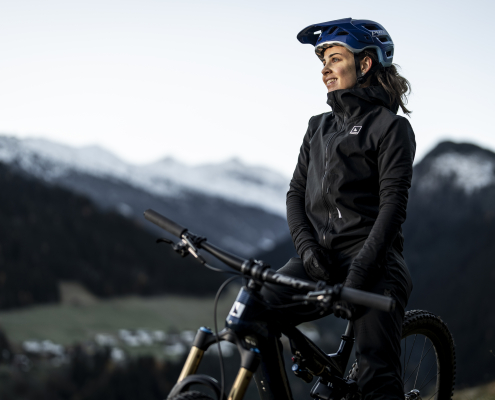 Eine Bikerin, mit blauem Helm, sitzt auf ihrem Bike und genießt die Aussicht. Eine beeindruckende Bergfront sowie ein kleines Tal bilden den Hintergund.
