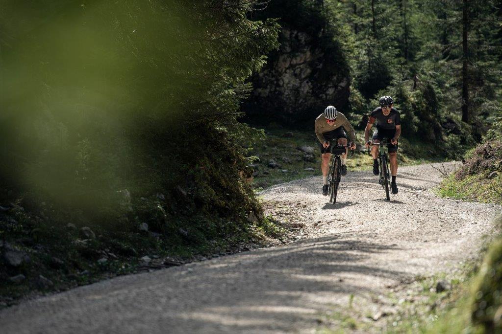 Zwei männliche Radfahrer fahren einen Forstweg hinauf. Der linke Fahrer trägt ein grünes langärmeliges Trikot, der andere hingegen ein kurzes schwarzes. Bei tragen Helme und Brillen.