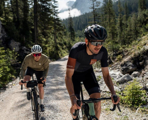 Zwei männliche Radfahrer fahren einen Forstweg hinauf. Der hintere Fahrer trägt ein grünes langärmeliges Trikot, der vordere hingegen ein kurzes schwarzes. Bei tragen Helme und Brillen. Im Hintergrund erstreckt sich eine Wald- und Berglandschaft.
