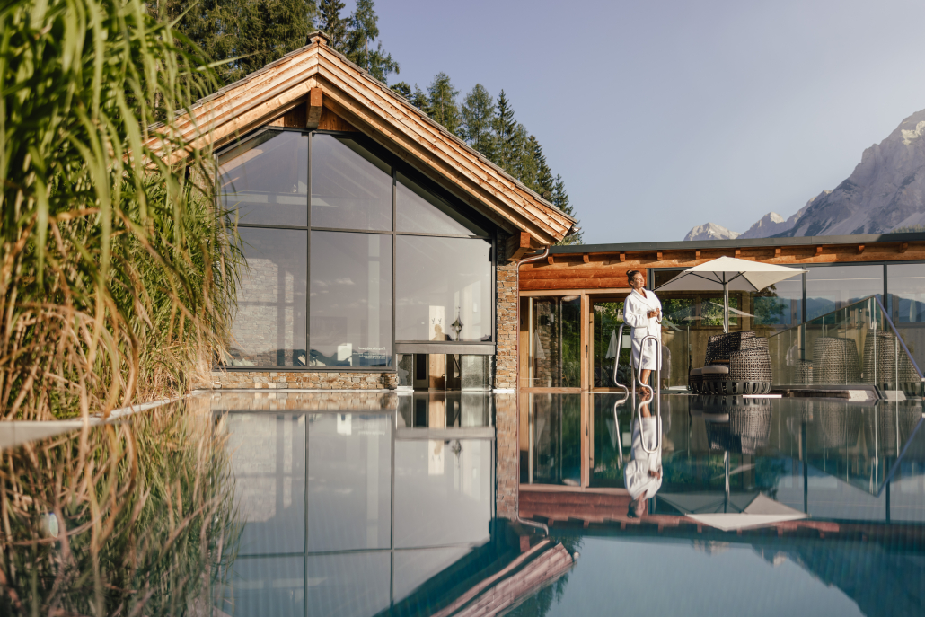 Eine Frau im Bademantel steht an einem Pool vor einem modernen Gebäude mit Glasfront, Bäumen und Bergen im Hintergrund.
