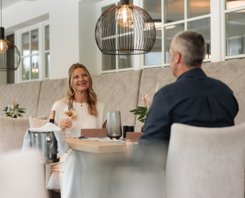 Eine Frau in einem weißen Kleid, mit einem Glas Weißwein in der rechten Hand, sitzt mit einem Mann in einem schwarzen Hemd an einem gedeckten Tisch. Dieser ist geschmückt mit einer Blumenvase sowie einem Teelichthalter. Auf der linken Seite des Tisches befindet sich ein Weinkühler, auf der anderen geht eine Sitzbank entlang. Im Hintergrund sind ebenfalls gedeckte Tische. Über jedem einzelnen Tisch ist jeweils ein großer Lampenschirm.