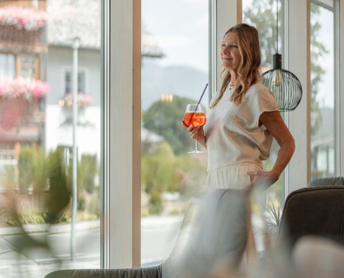 Eine Frau steht entspannt vor einer Fensterfront. In der Hand hält sie ein Glas Aperol-Spritz. Im Vordergrund befinden sich einige gemütlich aussehende Couchsesseln. Im Hintergrund sieht man durch die Fenster ein großes Haus mit Balkonen die voller Blumen sind und eine bergige Naturlandschaft.