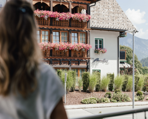 Das Bild zeigt eine Frau von hinten, die auf ein traditionelles Hotel mit Holzfassade und vielen blühenden Blumenkästen schaut. Im Hintergrund sind Berge und ein klarer Himmel zu sehen – eine ruhige, alpenländische Szenerie.