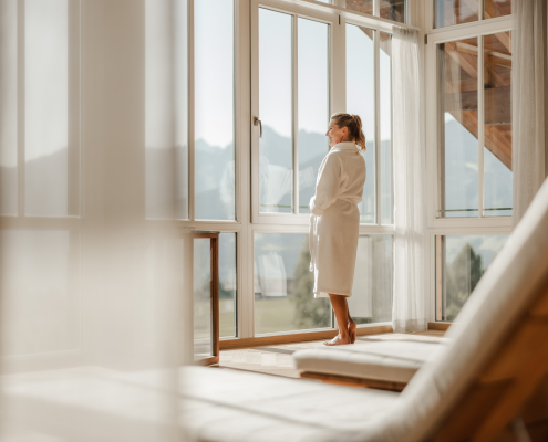 Im Fokus steht eine Frau im Bademantel die aus dem Fenster sieht und die Aussicht genießt. Im Vordergrund, aber verschwommen, sind Liegen zu erkennen.
