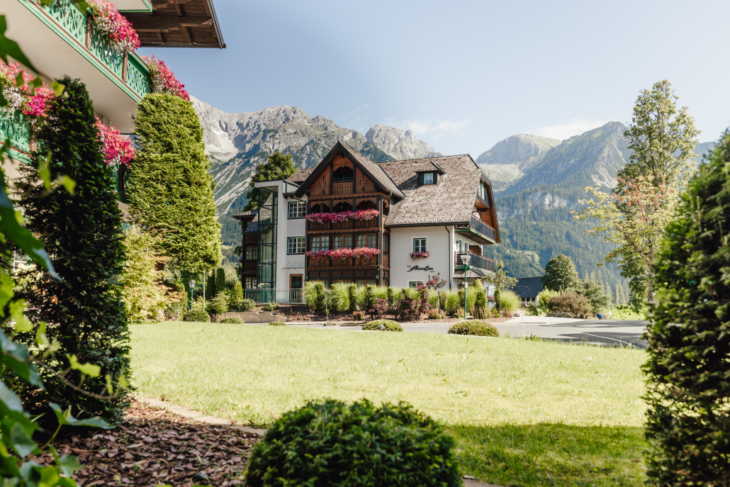 Das Bild zeigt ein traditionelles, mehrstöckiges Gebäude mit Holzbalkonen und vielen Blumenkästen, umgeben von gepflegtem Grün und hohen Bäumen. Im Hintergrund sind imposante Berge und viele Bäume zu sehen.
