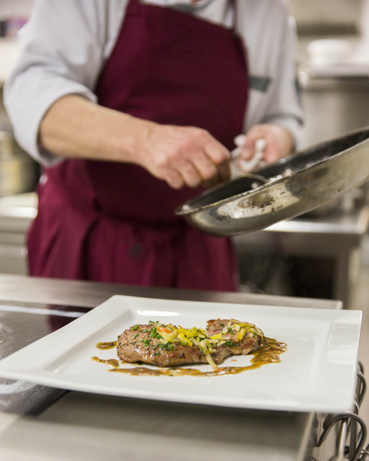 Ein Koch richtet ein Stück Fleisch auf einem Teller abetietlich an. Garniert mit einer Soße und feingehacktem Gemüse auf einem weißen Teller.