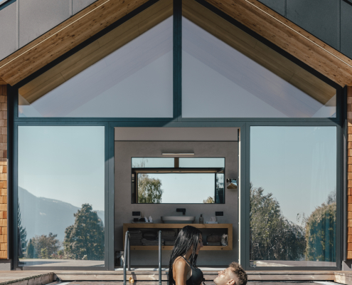 Ein Paar befindet sich in einem Außenpool vor einem modernen Haus mit großer Glasfront. Die Frau trägt einen schwarzen Bikini und wird vom Mann im Wasser gehalten. Im Hintergrund sind Bäume und Berge zu sehen.