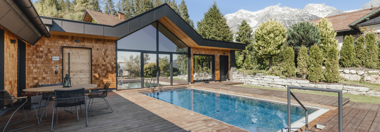 Ein modernes Haus mit Holzfassade und großen Glasfronten. Vor dem Haus befindet sich ein großer Außenpool. Eine Holzterrasse mit Sitzgelegenheiten umgibt den Pool. Im Hintergrund sind Bäume und schneebedeckte Berge zu sehen.