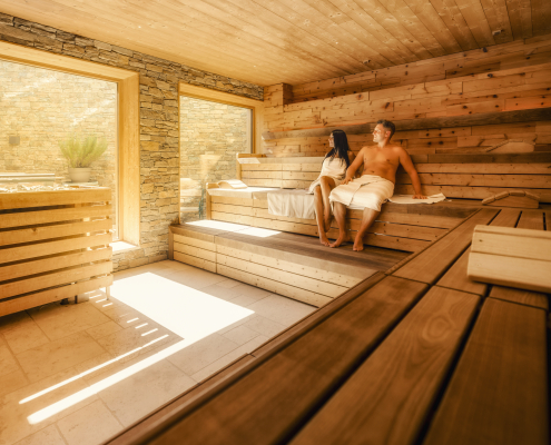 Ein Paar entspannt in einer großen modernen Sauna mit großen Fenstern und Holzbänken.