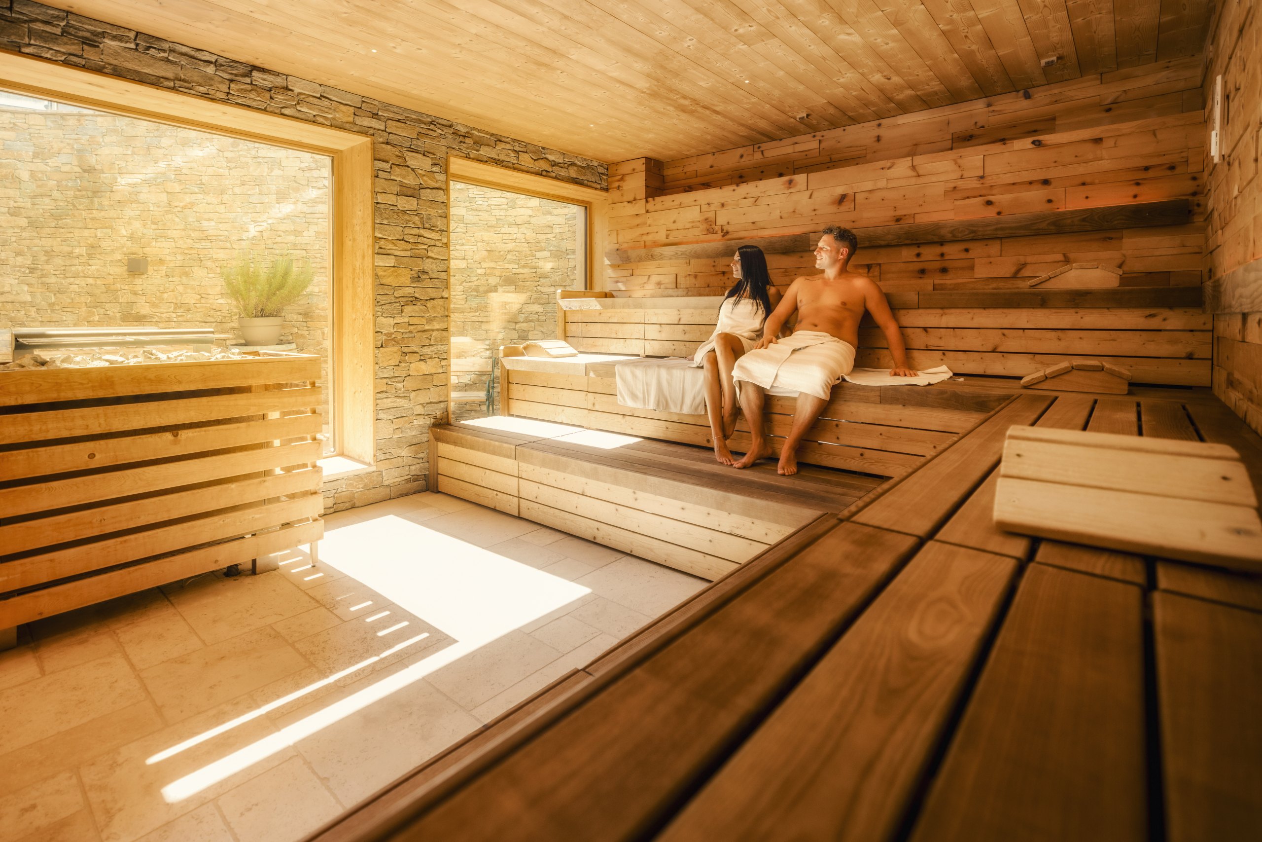 Ein Paar entspannt in einer großen modernen Sauna mit großen Fenstern und Holzbänken.