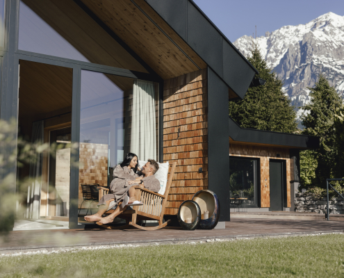 Ein Pärchen sitzt gemeinsam auf einem Schaukelstuhl, beide in Bademänteln. Die Frau sitzt auf dem Schoß des Mannes. Im Hintergrund steht ein modernes Haus mit großer Fensterfront, umgeben von Bäumen und Bergen