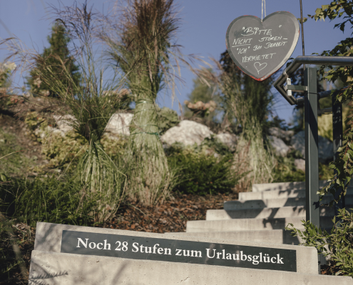 Im Vordergrund sind Betontreppen mit einem Schild " Noch 28 Stufen zum Urlaubsglück". Darüber, an einer Stange befestigt, hängt ein Schild in Form eines Herzens, auf dem alles groß geschrieben ist " Bitte nicht stören " Ich" bin derzeit verheiratet" , beendet wir diese Nachricht mit einem Herzzeichen. Die Treppen sind auf der rechten Seite begrenzt mit einem Geländer und auf der anderen Seite mit Blumen und höhen Gräsern.