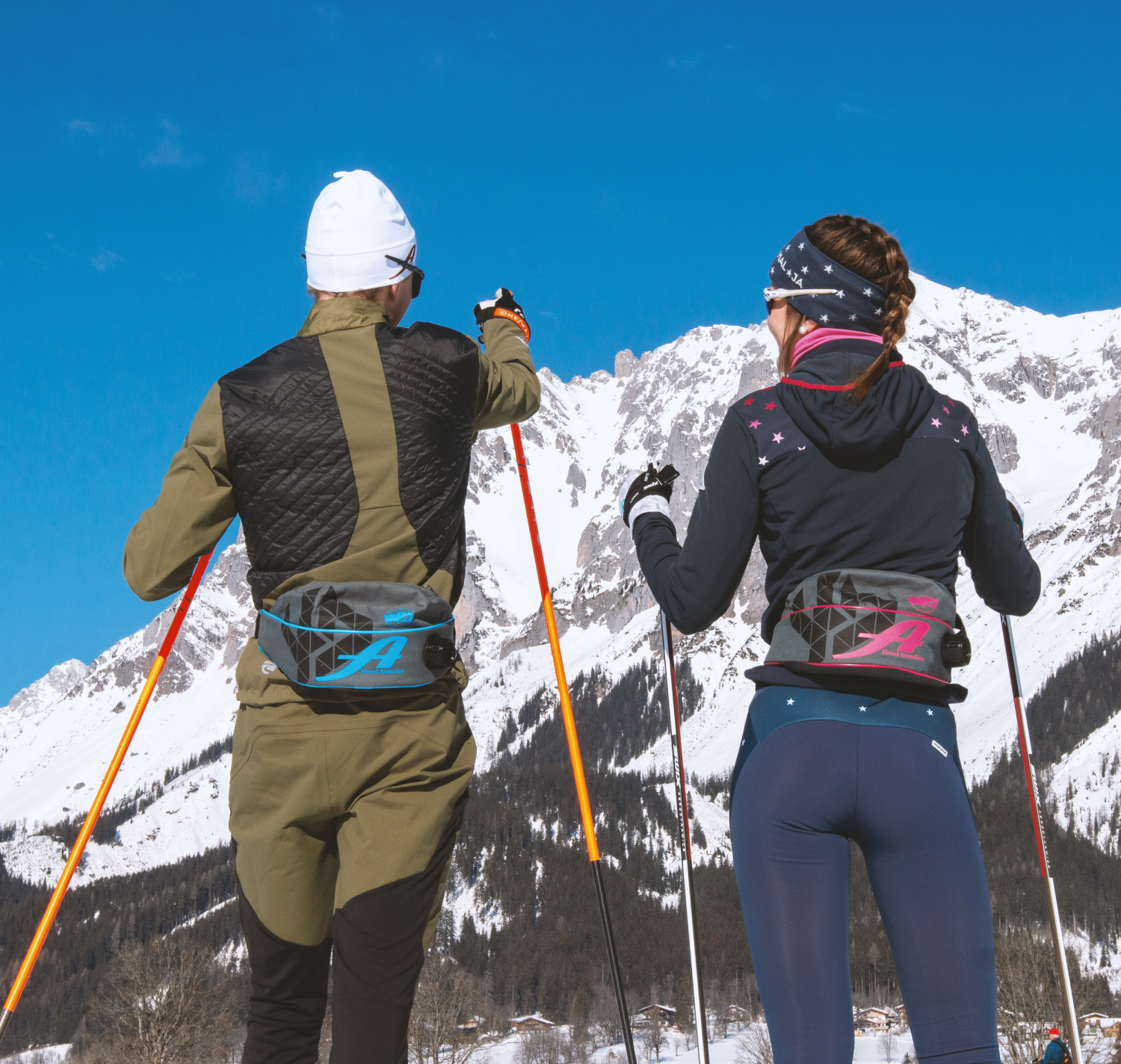 Zwei Menschen stehen mit Skistöcken im Schnee und blicken auf eine große, verschneite Berglandschaft unter blauem Himmel.