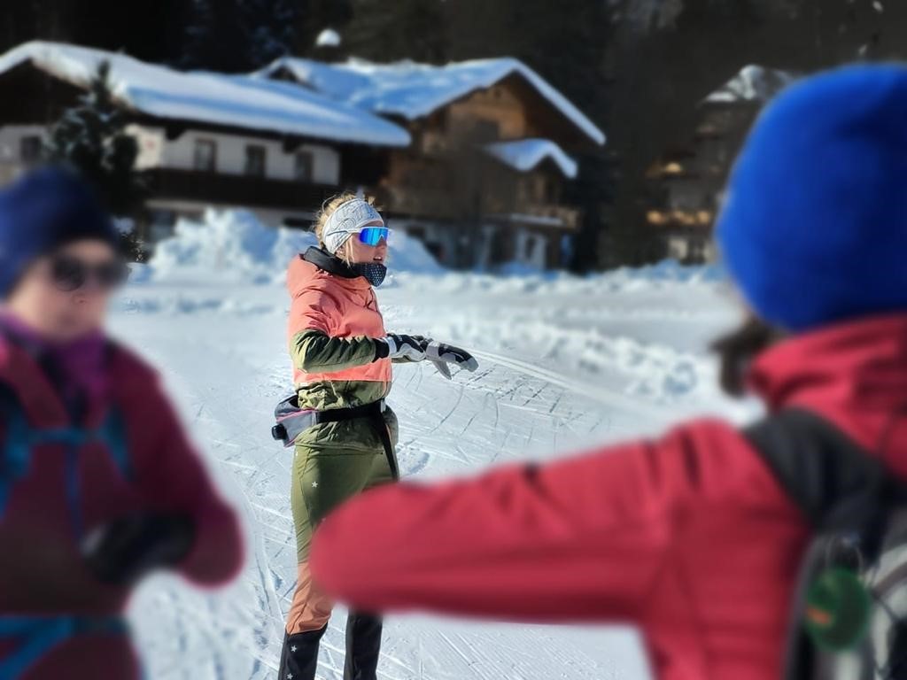 Skischule Ramsau Annelies Das Bild zeigt drei Menschen in Winterbekleidung, wobei eine Frau mit blondem Pferdeschwanz und Sonnenbrille in der Mitte steht und zu den anderen beiden blickt, die unscharf im Vordergrund sind. Im Hintergrund sind verschneite Häuser und Bäume zu erkennen.