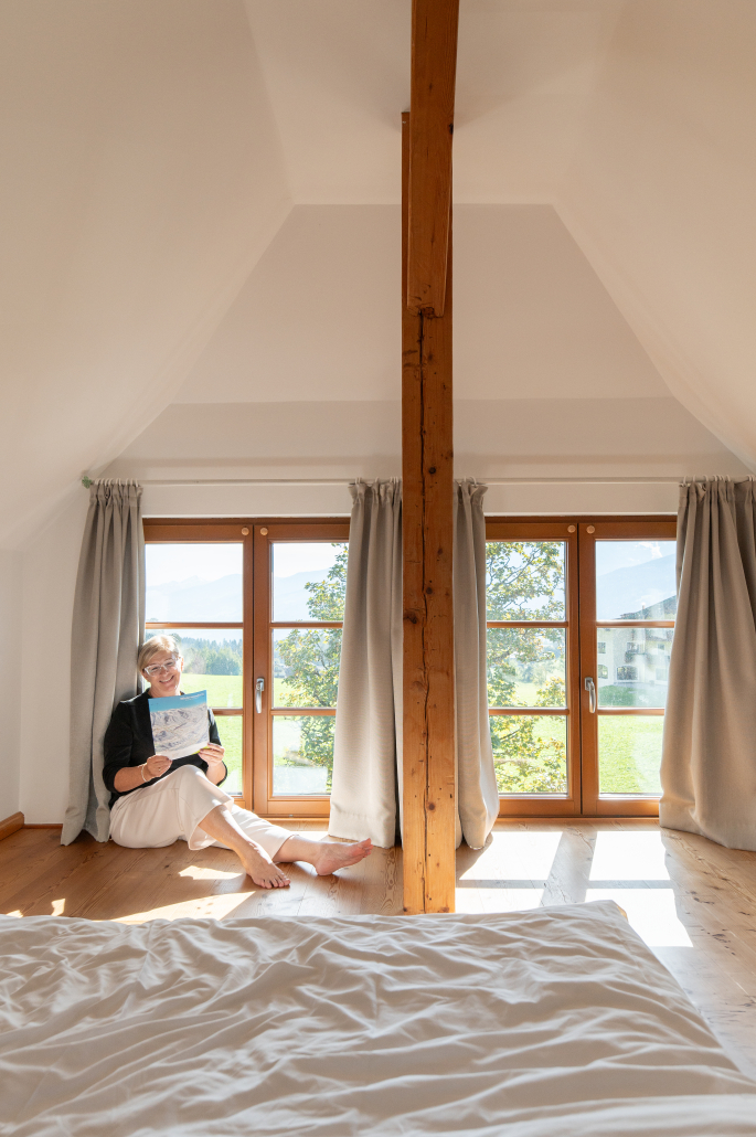 Frau in hellem Dachzimmer Eine Frau sitzt lesend vor großen Fenstern in einem hellen Dachzimmer mit Holzboden und Blick ins Grüne.