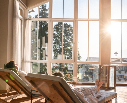 Zwei Personen entspannen auf Holzliegen in einem hellen Raum mit großen Fenstern, die viel Sonnenlicht hereinlassen und eine Aussicht ins Freie bieten.