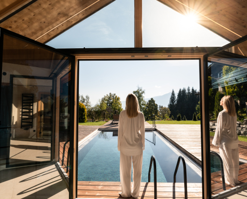 Eine Frau in weißer Kleidung steht in einem modernen Holzgebäude mit großen Glasfronten und blickt auf einen Außenpool und die dahinterliegende Landschaft mit Bergen.