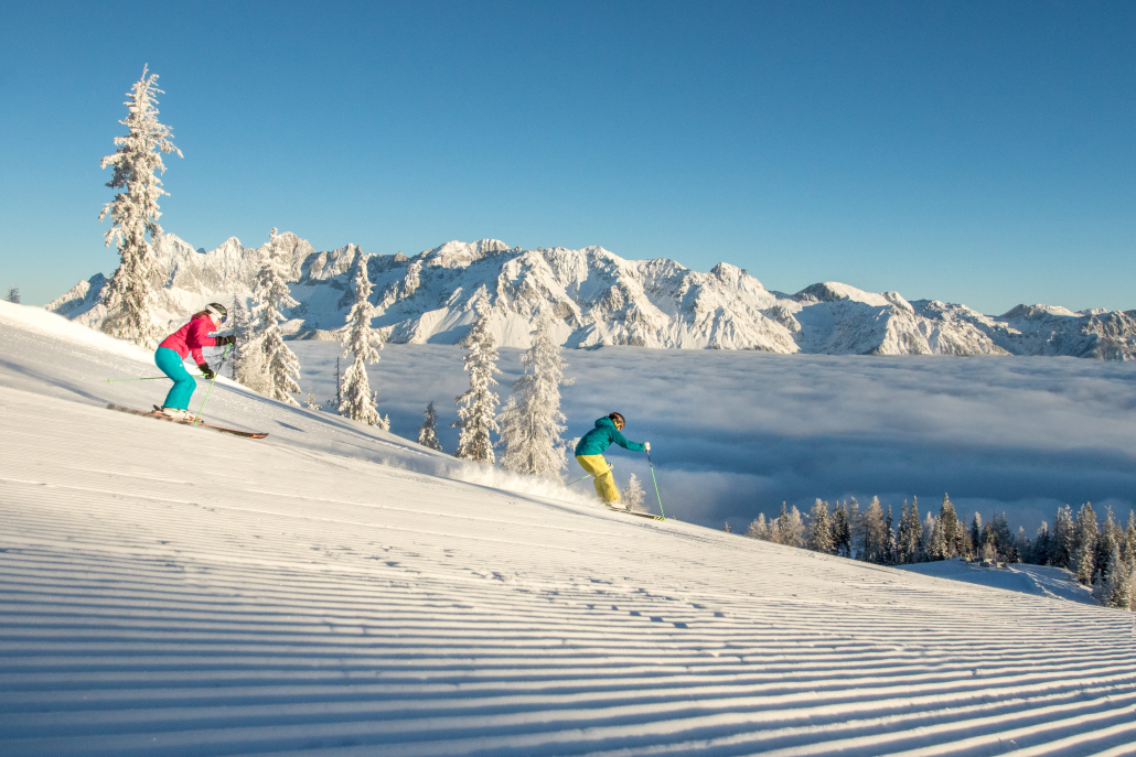 Zwei Skifahrer auf einer Piste, im Hintergrund eine alpine Winterlandschaft mit hohen Bergen.