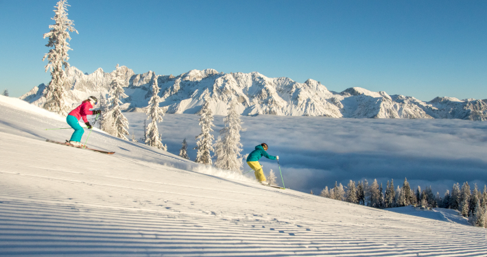 Skifahren Hochwurzen Zwei Skifahrer auf einer Piste, im Hintergrund eine alpine Winterlandschaft mit hohen Bergen.