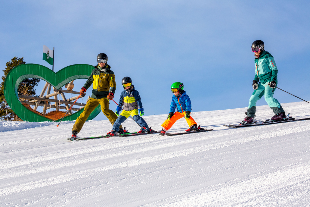 Zwei Kinder und zwei Erwachsene in Wintersportkleidung stehen mit Skiern auf einer Piste. Alle tragen Helme und Skibrillen, die Erwachsenen auch Skistöcke. Im Hintergrund ist ein großes grünes Herz mit der Steiermark-Flagge zu sehen. Der Himmel ist klar und blau.