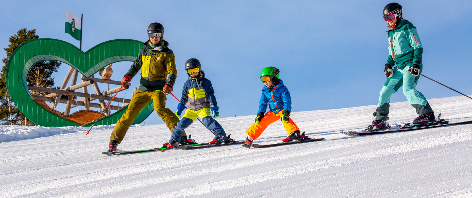 Skifahren Reiteralm Zwei Kinder und zwei Erwachsene in Wintersportkleidung stehen mit Skiern auf einer Piste. Alle tragen Helme und Skibrillen, die Erwachsenen auch Skistöcke. Im Hintergrund ist ein großes grünes Herz mit der Steiermark-Flagge zu sehen. Der Himmel ist klar und blau.