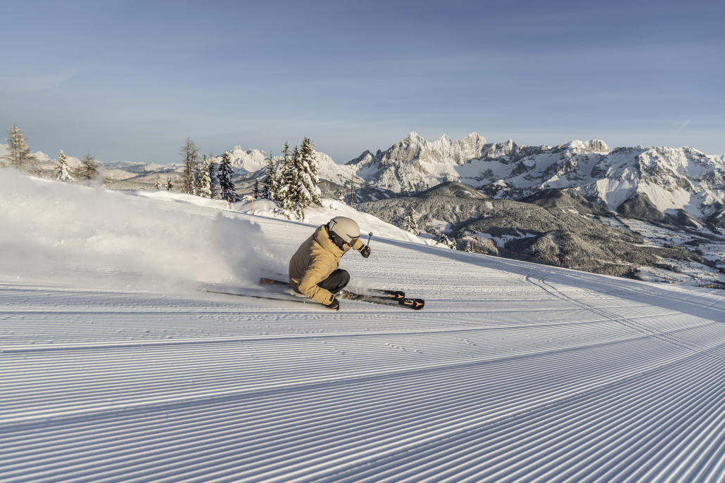 Eine Skifahrerin fährt auf einer Piste. im Hintergrund ist eine beeindruckende Bergkulisse zu sehen.