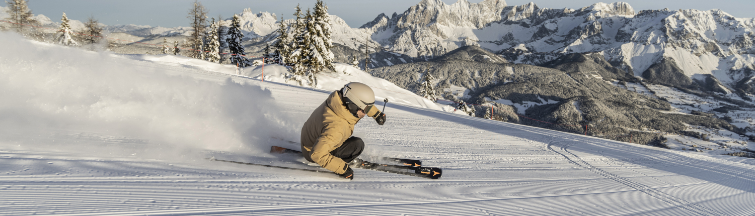 Eine Skifahrerin fährt auf einer Piste. im Hintergrund ist eine beeindruckende Bergkulisse zu sehen.