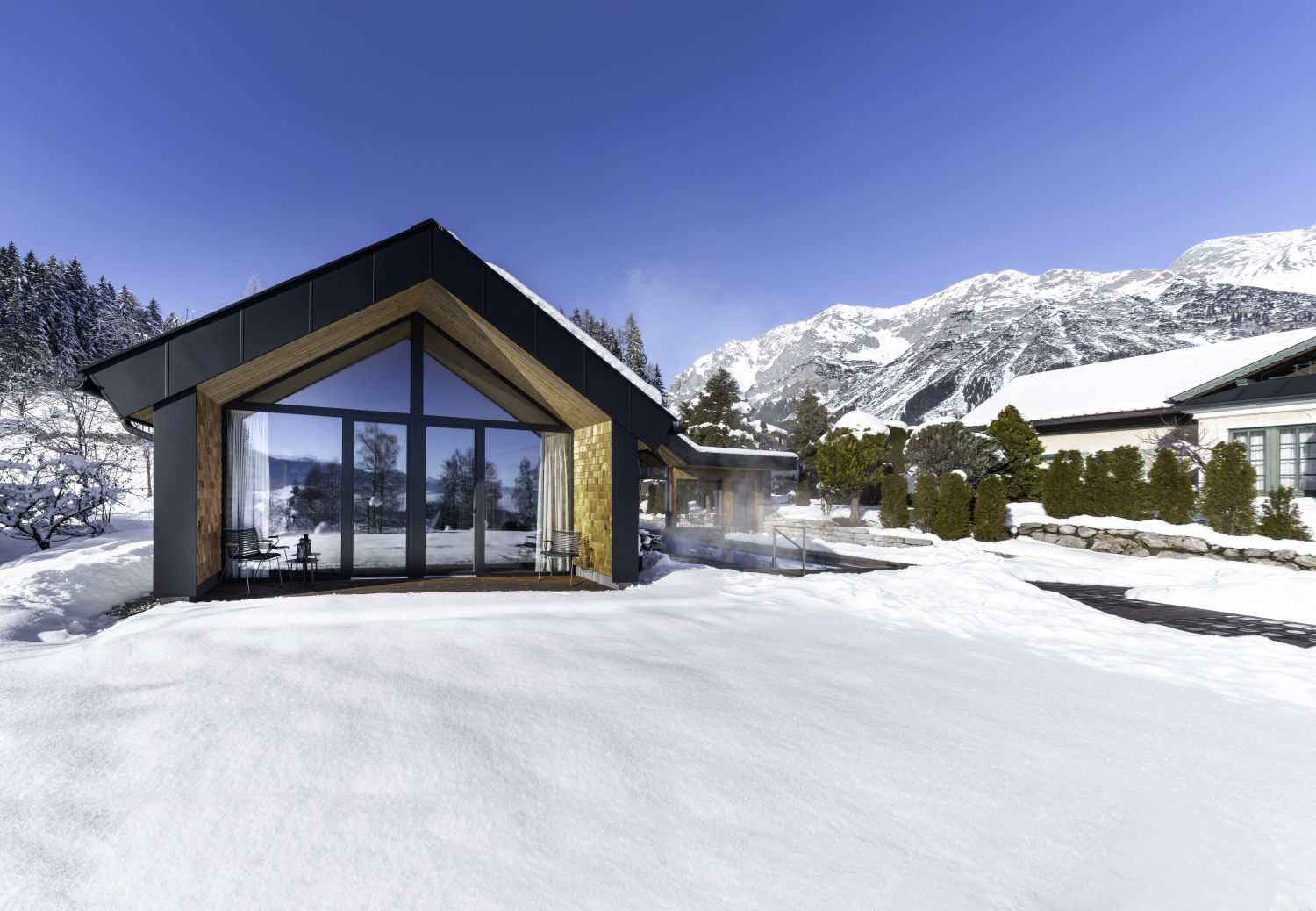 Zusehen ist ein modernes Holzhaus mit großen Fenstern in den verschneiten Bergen. Neben ihm befindet sich ein warmer Außenpool.