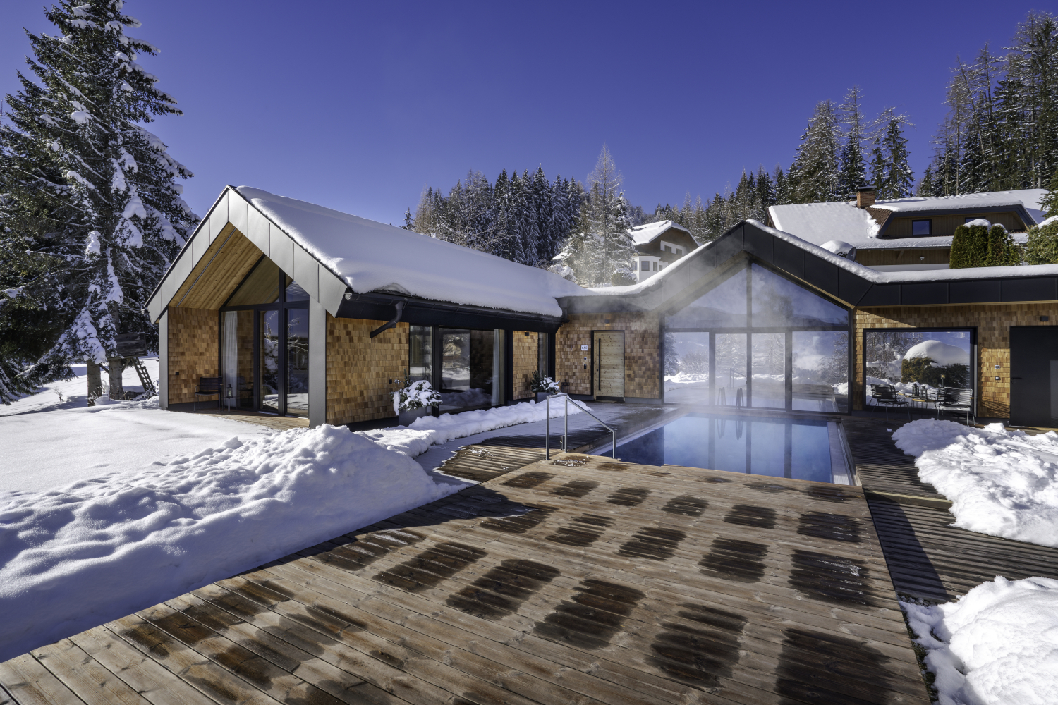 Bild zeigt ein modernes Holzhaus in den verschneiten Bergen. Vor dem Haus liegt eine Holzterasse mit einem warmen Außenpool. Um das Haus liegt viel Schnee, und im Hintergrund stehen viele verschneidte Bäume unter blauem Himmel.