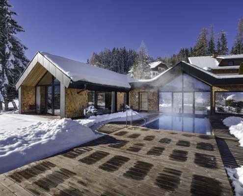 Bild zeigt ein modernes Holzhaus in den verschneiten Bergen. Vor dem Haus liegt eine Holzterasse mit einem warmen Außenpool. Um das Haus liegt viel Schnee, und im Hintergrund stehen viele verschneidte Bäume unter blauem Himmel.