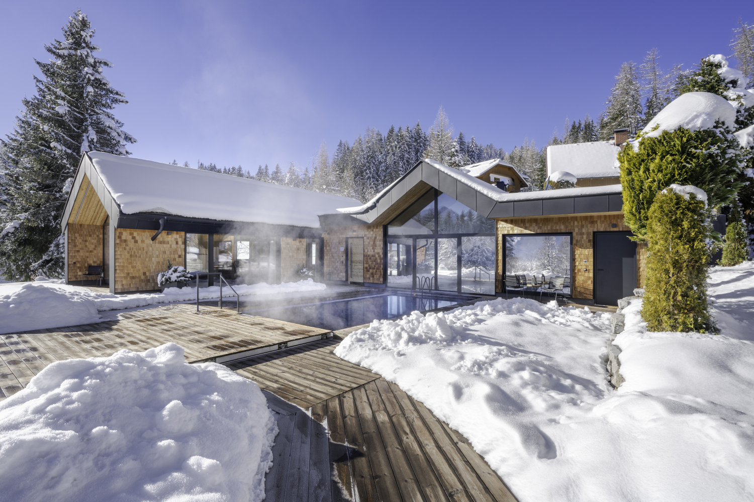 Ansicht des Luxus Chalet vom Holzweg, der zum Haus führt. Zusehen ist der Pool und mehrere rießige Fenster.