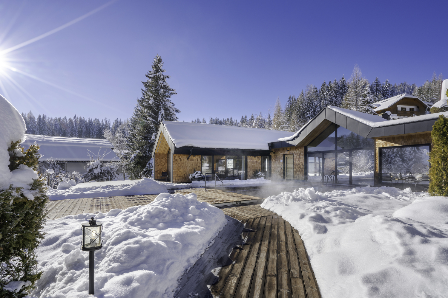 Ansicht des Luxus Chalet vom Holzweg, der zum Haus führt. Zusehen ist ein warmer Außenpool und mehrere rießige Fenster. Sowie im Hintergrund viele verschneidte Bäume.