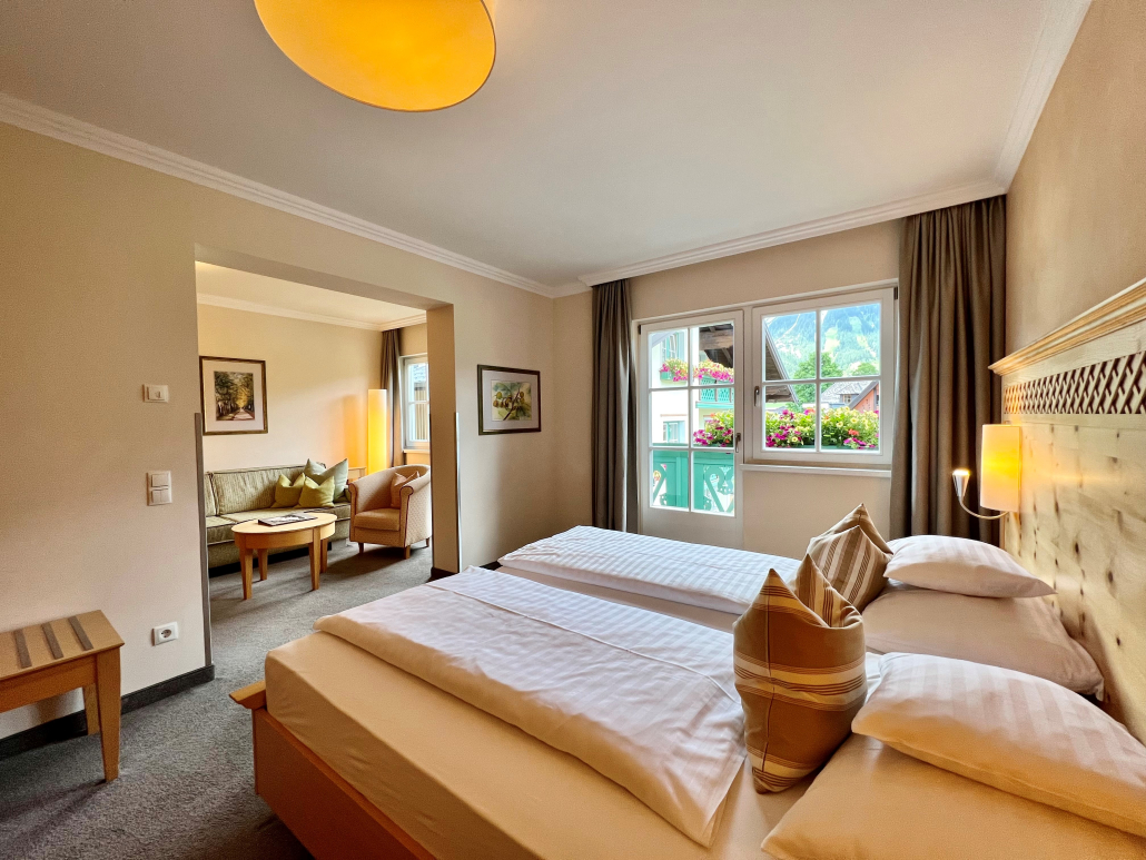 Das Bild zeigt das Schlafzimmer einer Juniorsuite mit Balkon und dem gegenüberliegenden Wohnbereich mit Sofa, Sessel und Couchtisch.